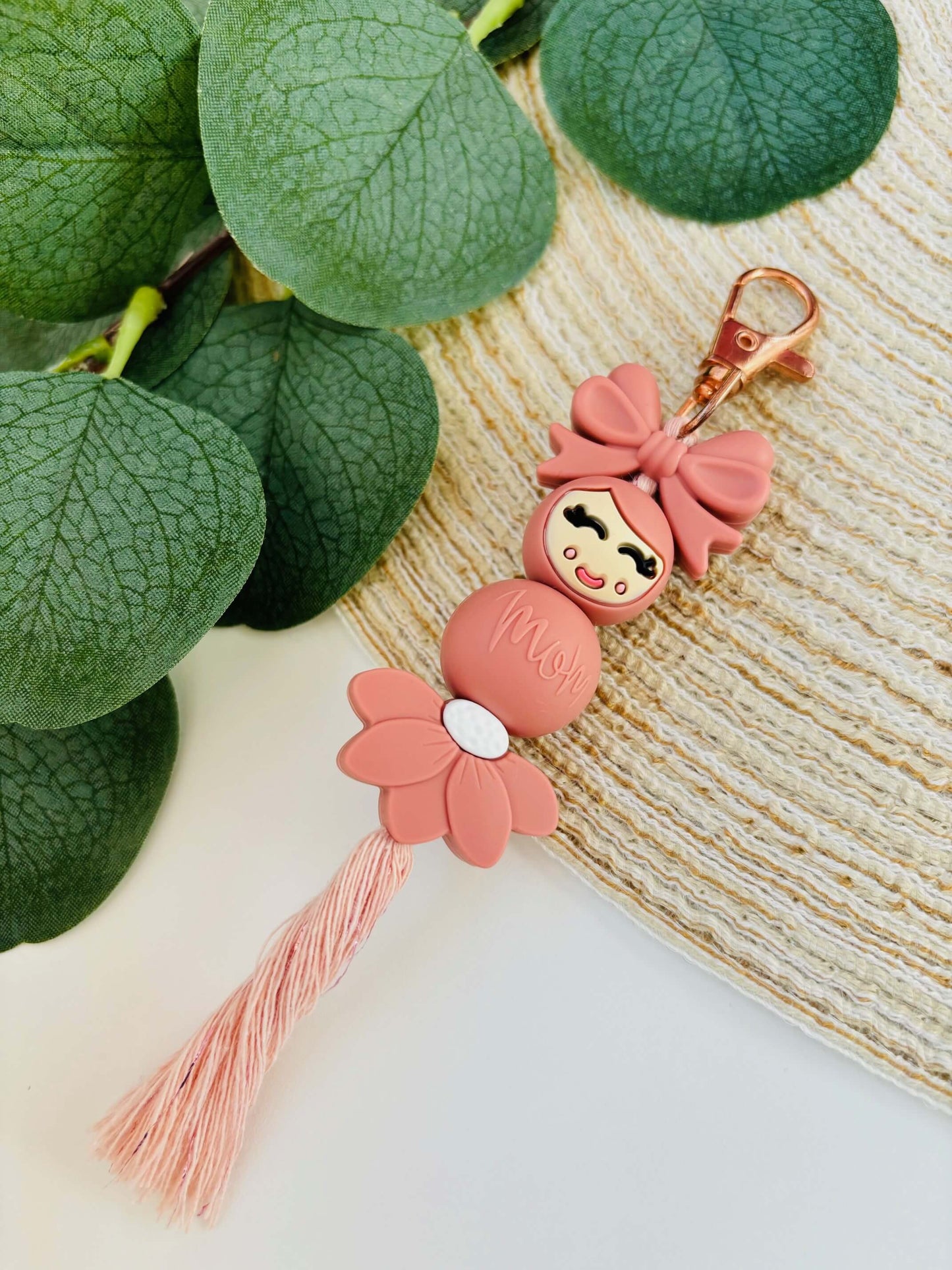 Handgemaakte siliconen popje van geluk in roze kleur. Het strikje en bloemetje maken het extra vrouwelijk, gecombineerd met een vleugje macramé.