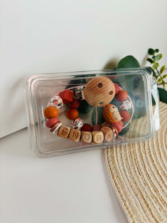 Handgemaakte speenkoord en bijtring met naam gemaakt van siliconen kralen in warme herfst kleuren rood, oranje en hout met een printkraal met bloemenmotief. Verpakt in een transparant doosje.