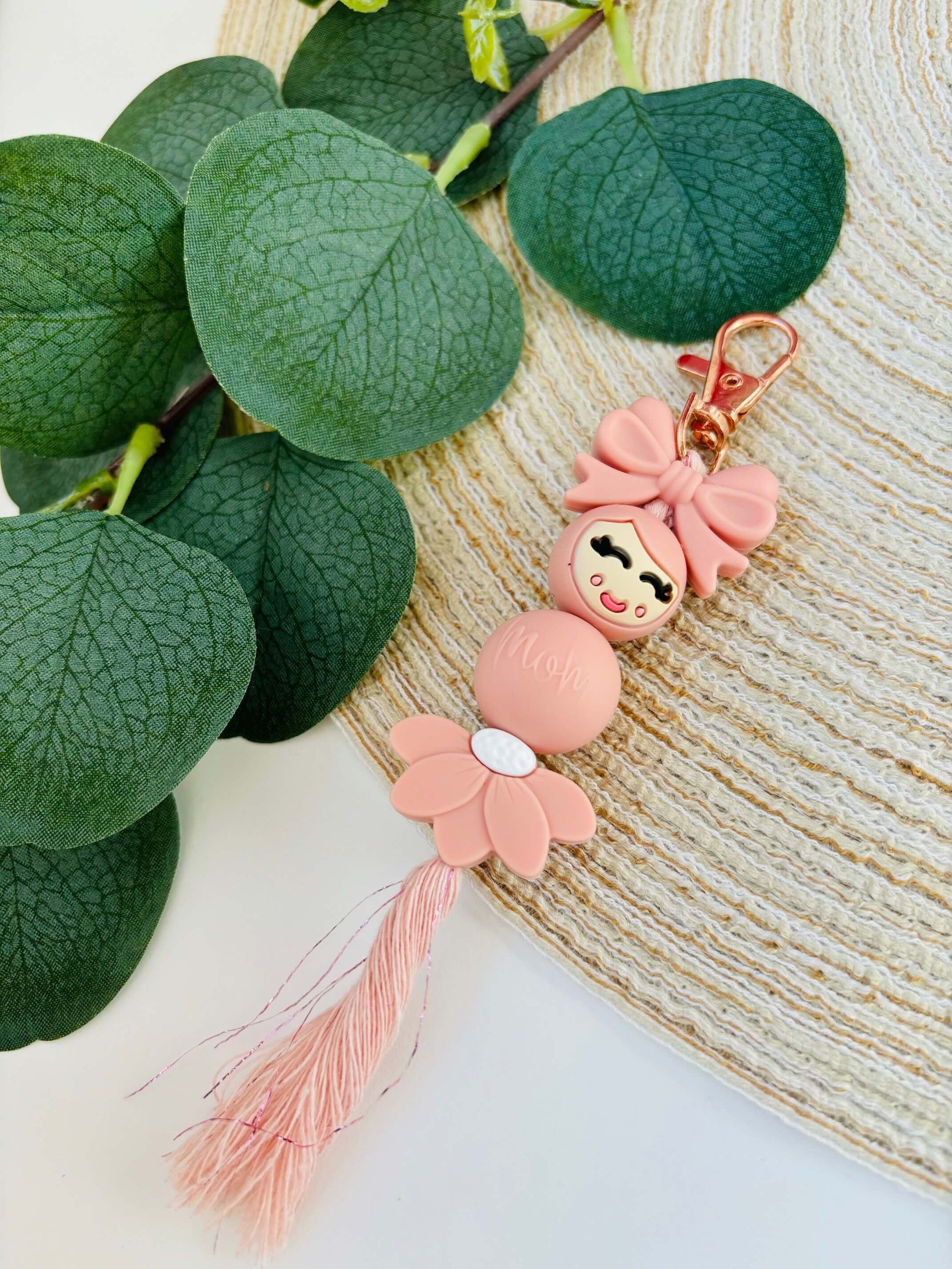 Handgemaakte siliconen popje van geluk in roze kleur. Het strikje en bloemetje maken het extra vrouwelijk, gecombineerd met een vleugje macramé.